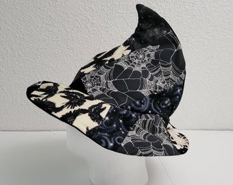 May include: A black and white witch hat with a pointed crown and wide brim. The hat features a patchwork design with raven, spiderweb, and celestial patterns. The hat is displayed on a white mannequin head.