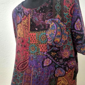 Könnte beinhalten: Langärmliges Oberteil mit einem Patchwork-Design mit Paisley-Mustern in Lila, Rot, Türkis und Gold. Das Oberteil hat einen runden Ausschnitt und einen dunklen Hintergrund. Der Stoff scheint weich und strukturiert zu sein.