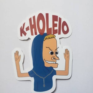 May include: A die-cut sticker featuring a cartoon character in a blue hooded outfit with the text "K-HOLEIO" in red letters. The character has a distinctive facial expression and raised arms.