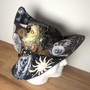 May include: A pointed fabric hat featuring a patchwork design with black, white, and gold celestial motifs. The pattern includes illustrations of a crescent moon with a face, a sun with a face, stars, constellations, and a skull on a dark background.