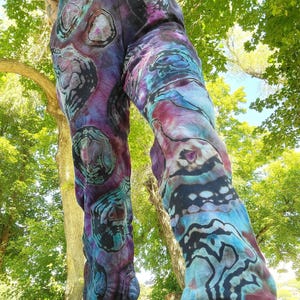 Pode incluir: Calças de moletom tie-dye em tons de roxo, azul e preto. As calças apresentam um padrão único com formas orgânicas e cós com cordão. A parte inferior das calças é canelada.