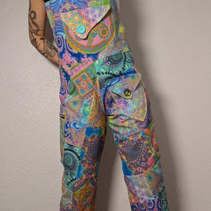 May include: A colorful jumpsuit with a patchwork design featuring geometric and floral patterns in shades of blue, pink, green, and yellow. The jumpsuit has two front pockets with button closures and a black top.