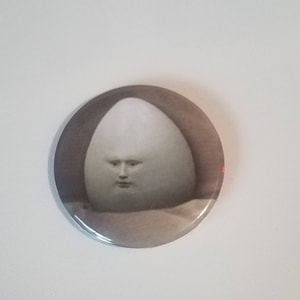 May include: A white egg with a human face on it. The egg is on a brown background.