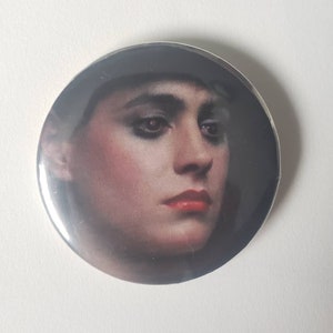 May include: A close-up of a round button featuring a black and white photo of a woman with dark hair and dark eye makeup. Her lips are painted a bright red.