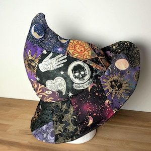 May include: A pointed hat made of patchwork fabric featuring celestial and mystical motifs including suns, moons, stars, skulls, and a palmistry hand. The fabric displays a dark color palette of black, purple, and gold with the text So Below.
