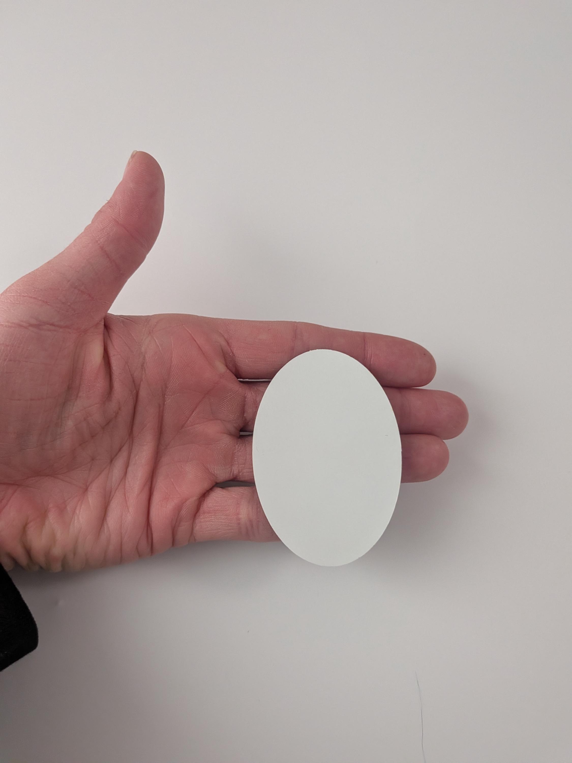 Puede incluir: Un objeto blanco de forma ovalada. El objeto se sostiene en la mano de una persona sobre un fondo blanco. La superficie lisa del objeto refleja la luz. La forma es similar a un huevo o un rect&aacute;ngulo redondeado.
