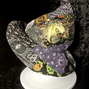 May include: A patchwork witch hat featuring a variety of printed fabric panels. The patterns include a crescent moon with flowers, skeletons, snakes, stars, and green skulls on a black background. The hat has a wide brim and a pointed crown.