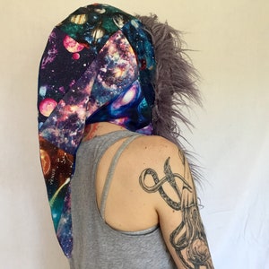 Op de afbeelding: Een kledingstuk met capuchon met een galaxy print en een paarse nep bont trim. De capuchon is gevormd als een puntige hoed en heeft een lange staart. De print bevat planeten, sterren en galaxies. Het kledingstuk is gemaakt van een zachte, comfortabele stof.