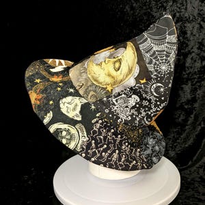 May include: A pointed hat featuring a patchwork design of black fabric panels with various white and gold illustrations, including a crescent moon with a face, spider webs, skeletons, skulls, and stars, constructed with a wide, curved brim.
