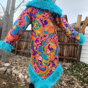 May include: A long, turquoise faux fur-trimmed coat with a colourful paisley print. The coat has a large collar and a long, flowing silhouette.