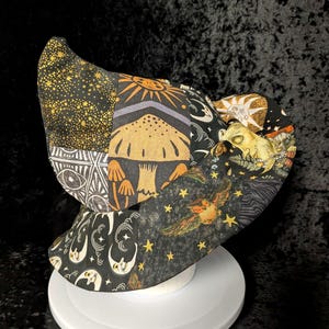 May include: A patchwork bonnet featuring a variety of black fabric panels with gold and white printed designs including mushrooms, suns, crescent moons, stars, an owl, and geometric eye patterns. The bonnet has a wide, curved brim and a structured crown.