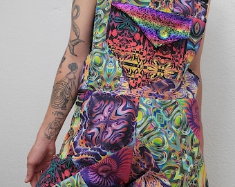 May include: A colorful romper with a psychedelic pattern. The romper features a patchwork design with vibrant hues of pink, purple, green, and yellow. It has adjustable black straps and a front pocket with a rainbow iridescent finish. The garment is a one-piece outfit.