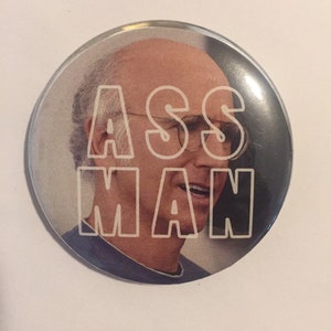 Op de afbeelding: Een ronde knop met een witte omtrek van de woorden "ASS MAN" op een foto van een man met een kale kop en een bril.