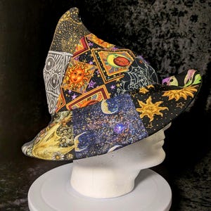 May include: A patchwork bucket hat featuring a variety of celestial and mystical patterns, including suns, moons, stars, and spiderwebs in shades of black, gold, yellow, and purple. The hat has a pointed crown and a wide, downward-sloping brim.