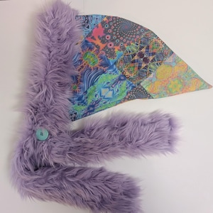 May include: A lavender-colored, furry scarf with a large, colorful, patterned fabric accent. The scarf has a button closure and a soft, plush texture. The fabric accent features a mix of geometric and abstract designs.