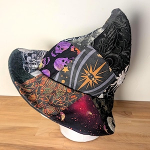 May include: A patchwork witch hat constructed from various fabric panels featuring black, purple, and orange colors. The patterns include skulls, a central eye symbol, celestial moons, stars, and intricate floral motifs across the conical crown.