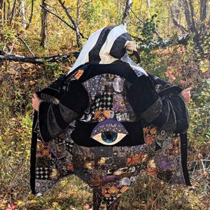 Può includere: Un mantello nero e fantasia con cappuccio, con un disegno a occhio sul davanti. Il mantello presenta vari motivi celesti e geometrici nelle tonalità del nero, viola e arancione. Il cappuccio è a righe bianche e nere.