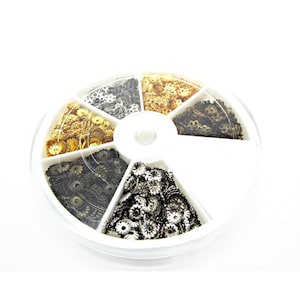 May include: A white plastic rotary storage container with six compartments filled with various sizes and colours of metal flower-shaped beads. The beads are gold, silver, and black.