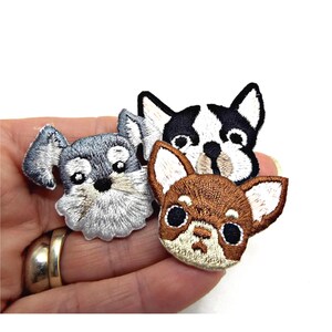 May include: Embroidered patches of three dog faces: a grey Schnauzer, a black and white Boston Terrier, and a brown Chihuahua. Each patch has detailed stitching and a white border, ideal for customising clothing or accessories.
