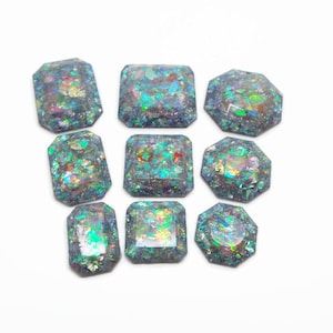 May include: A collection of eight faceted cabochons in rectangular, square, and octagonal shapes. Each piece features a dark base embedded with iridescent, multi-coloured foil flakes that reflect green, blue, pink, and gold light across the surface.