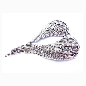 May include: A silver-coloured wing pendant with detailed feathering. The metal pendant is shaped like two wings arranged in a heart. It has a small loop at the top for hanging. The background is plain white, highlighting the pendant's intricate design.