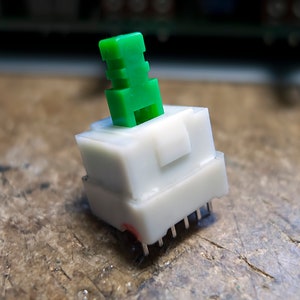 Op de afbeelding: Een witte mechanische toetsenbord schakelaar met een groene toetsdop. De schakelaar heeft acht pinnen aan de onderkant en is ontworpen voor gebruik in een toetsenbord.