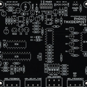 Puede incluir: Una placa de circuito impreso en blanco y negro con varios componentes electrónicos, incluyendo resistencias, condensadores, circuitos integrados y potenciómetros. La placa está etiquetada como "PHONIC TAXIDERMIST 47.2b" e incluye el texto "circuitbenders.co.uk".