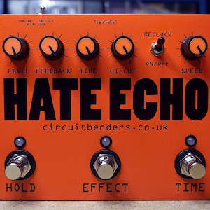 Puede incluir: Un pedal de efectos para guitarra de color naranja con el texto HATE ECHO en letras negras grandes. Cuenta con seis perillas con tapa naranja etiquetadas como Level, Feedback, Time, Hi-Cut y Speed, un interruptor Reclock y tres interruptores de pie.