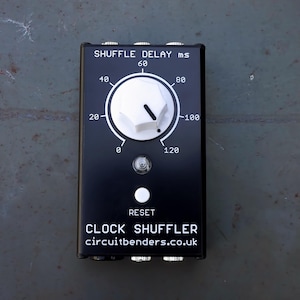 Pode incluir: Pedal preto Clock Shuffler com um botão branco e texto que diz "Shuffle Delay ms" e "Clock Shuffler". O pedal tem um botão de reset branco e duas tomadas de entrada/saída.