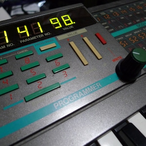 Può includere: Un sintetizzatore argento e nero con una manopola verde, un display digitale che mostra "33 97 98", e il testo "PROGRAM NO.", "PARAMETER NO.", "PROG/PARA", "VALUE", "VALUE", "VERIFY", "CANCEL", "SAVE", "LOAD", "WRITE", e "PROGRAMMER".