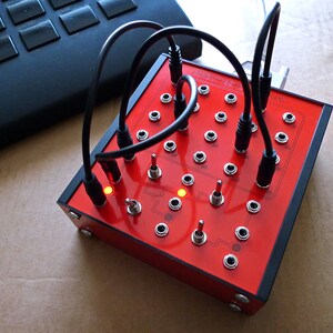 Puede incluir: Un dispositivo electrónico rojo con numerosos interruptores basculantes y puertos de conexión. Cables negros están enchufados al dispositivo. El dispositivo tiene un borde negro y está junto a un teclado negro.