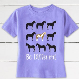 May include: A lilac-colored t-shirt featuring a design of nine horse silhouettes. Eight horses are black, and one is a brown and white pinto. The words "Be Different" are printed in white below the horses. The shirt has short sleeves and a crew neck.