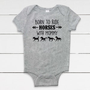 May include: A gray baby onesie with the text "BORN TO RIDE HORSES WITH MOMMY" in black. Below the text are four black silhouettes of horses running. The onesie is made of a soft material.