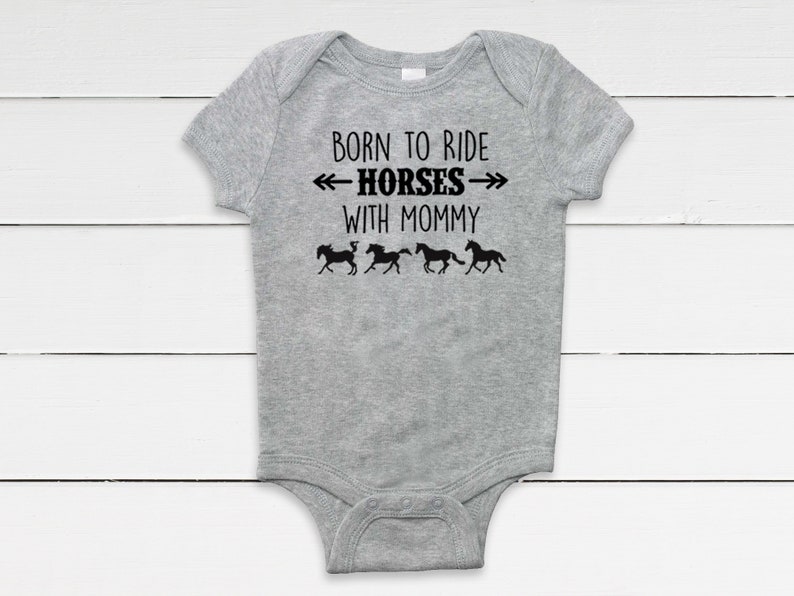 May include: A gray baby onesie with the text "BORN TO RIDE HORSES WITH MOMMY" in black. Below the text are four black silhouettes of horses running. The onesie is made of a soft material.
