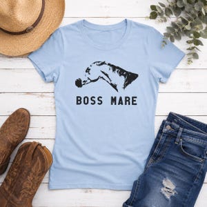 May include: Light blue t-shirt with a black horse head graphic and the text "BOSS MARE". The shirt is displayed with a straw hat, brown cowboy boots, and blue jeans on a white wooden surface.