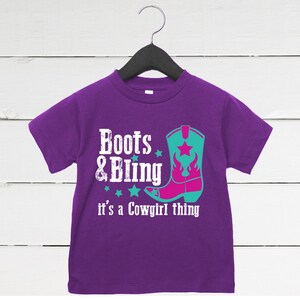 May include: A purple toddler t-shirt with the words "Boots & Bling" and "it's a Cowgirl thing" in white text. A turquoise and pink cowboy boot graphic is also featured on the shirt.