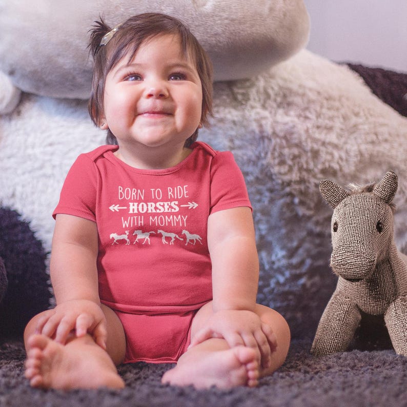 May include: A smiling baby wearing a red onesie with the text "Born to Ride Horses with Mommy" and horse silhouettes. A stuffed horse toy sits nearby. The baby is sitting on a gray carpet.