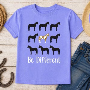 May include: A light purple t-shirt with a horse graphic and the text "Be Different". The graphic features eight black horses and one brown and white horse. A pair of blue jeans and a straw hat are also in the image.