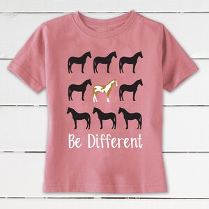 May include: A dusty rose t-shirt featuring a graphic of nine horse silhouettes. Eight are black, and one is a unique brown and white color. The text "Be Different" is printed in white below the horses.