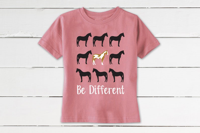 May include: A dusty rose t-shirt featuring a graphic of nine horse silhouettes. Eight are black, and one is a unique brown and white color. The text "Be Different" is printed in white below the horses.