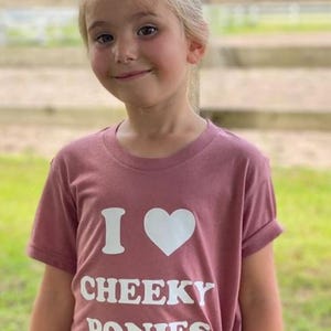 May include: A young person wearing a mauve t-shirt with the text "I heart Cheeky Ponies" in white.