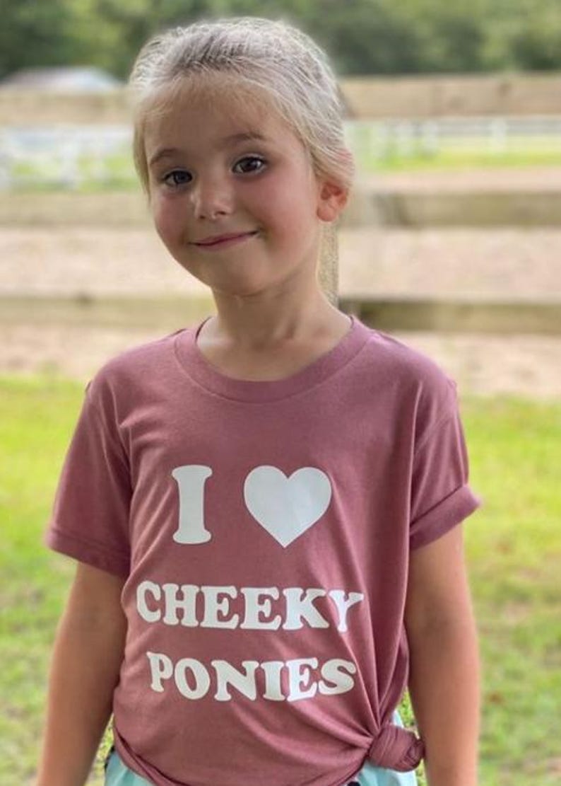 May include: A young person wearing a mauve t-shirt with the text "I heart Cheeky Ponies" in white.