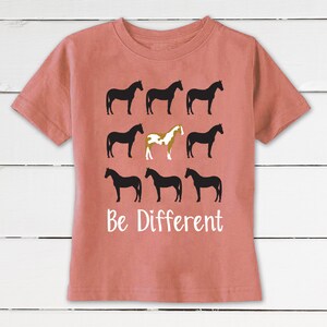 May include: A coral-colored t-shirt featuring a design of nine horse silhouettes. Eight horses are black, and one is a brown and white pinto. The words "Be Different" are printed in white below the horses.
