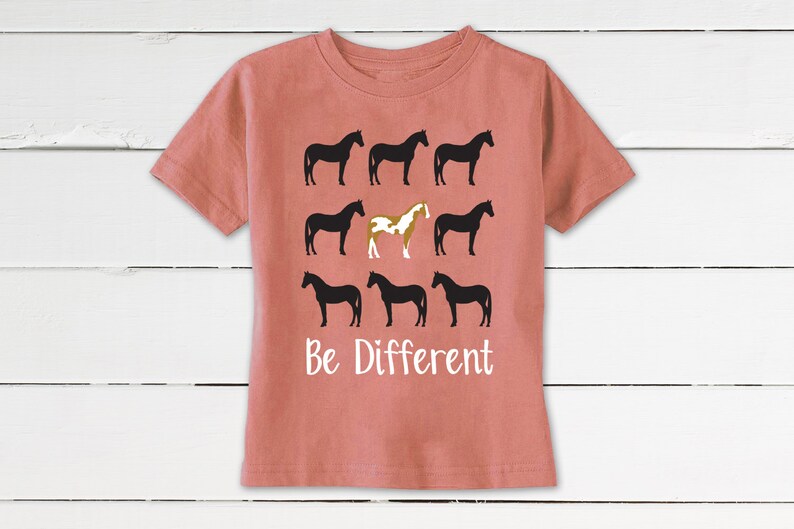 May include: A coral-colored t-shirt featuring a design of nine horse silhouettes. Eight horses are black, and one is a brown and white pinto. The words "Be Different" are printed in white below the horses.
