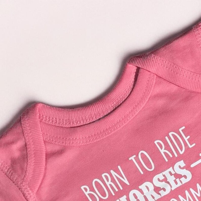 May include: Close-up of a pink baby onesie with the text "BORN TO RIDE HORSES". The onesie has a round neckline and short sleeves. The fabric is soft and appears to be cotton.