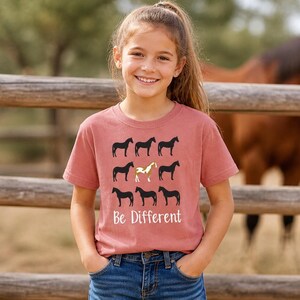 May include: A dusty rose t-shirt with a horse-themed design. Nine black horse silhouettes are arranged in a grid, with one gold horse in the center. The words "Be Different" are printed below the horses.