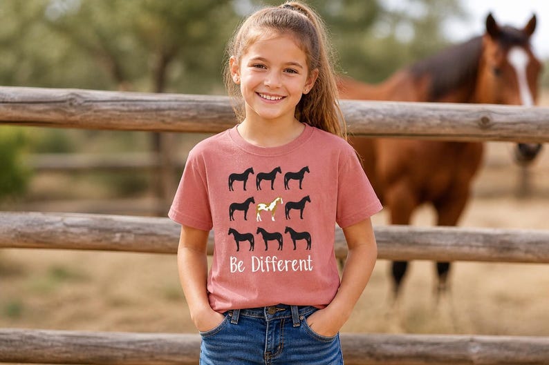 May include: A dusty rose t-shirt with a horse-themed design. Nine black horse silhouettes are arranged in a grid, with one gold horse in the center. The words "Be Different" are printed below the horses.