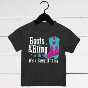 May include: Black t-shirt with the text "Boots & Bling" and "it's a Cowgirl thing". The shirt features a turquoise and pink cowboy boot graphic with a star and flame design. The shirt is hanging on a black hanger.