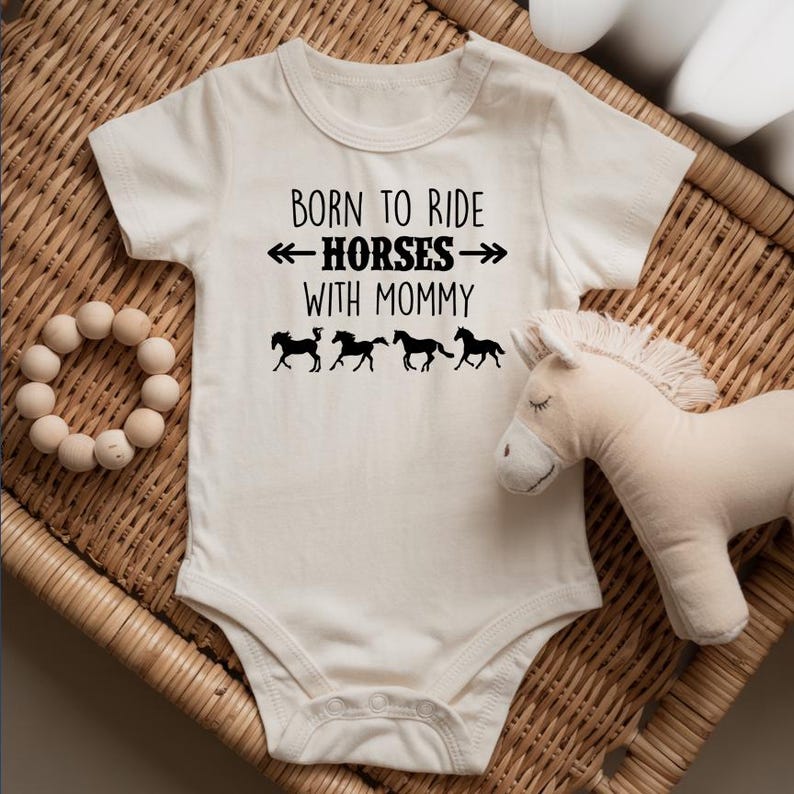 May include: Cream-colored baby bodysuit with the text "BORN TO RIDE HORSES WITH MOMMY" in black. Below the text are four black horse silhouettes. A wooden bead toy and a stuffed horse are also in the image.