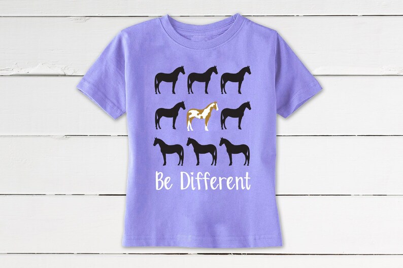 May include: A lilac-colored t-shirt featuring a design of nine horse silhouettes. Eight horses are black, and one is a brown and white pinto. The words "Be Different" are printed in white below the horses. The shirt has short sleeves and a crew neck.